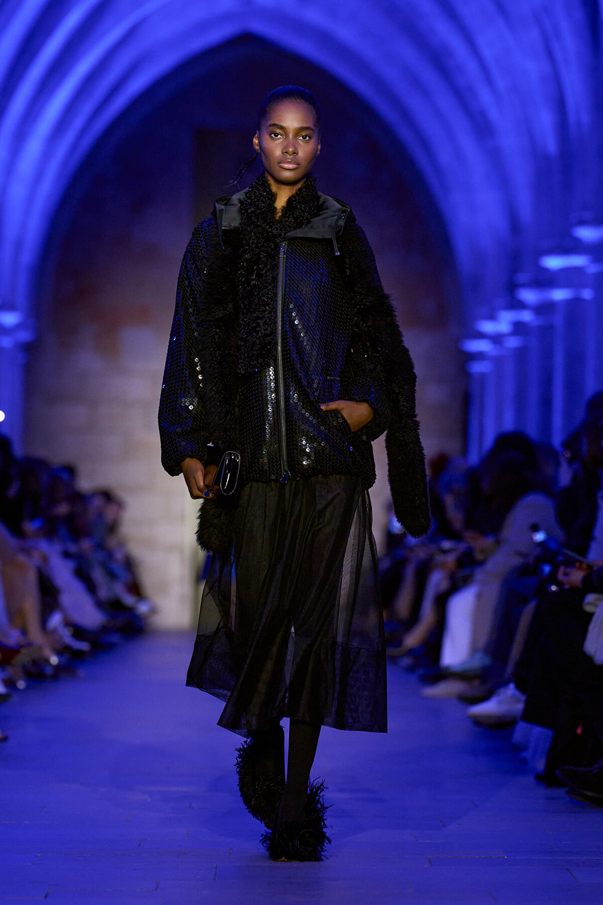 Model walking a fashion runway in a black sequined jacket, sheer black skirt, and feathered boots under blue arched lighting.