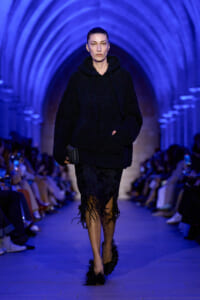 Model walking a blue-lit runway in a black fuzzy hoodie dress with a fringed hexagonal black skirt and small clutch.