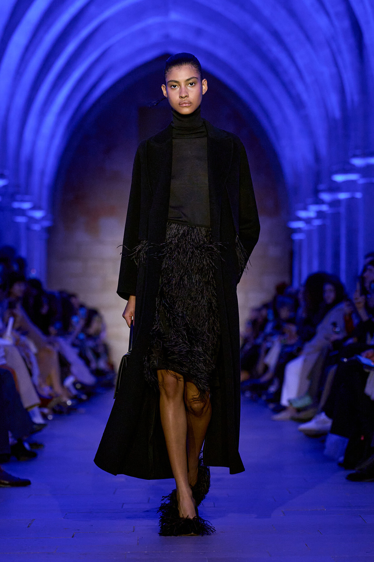 Model walking a runway in an all-black outfit—a turtleneck, long coat, and feathered skirt—under blue-illuminated arched vaults in a fashion show setting.
