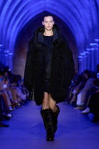 Model strides down a runway in a oversized black faux-fur coat, paired with fur boots, under blue-lit arches with an audience on sides