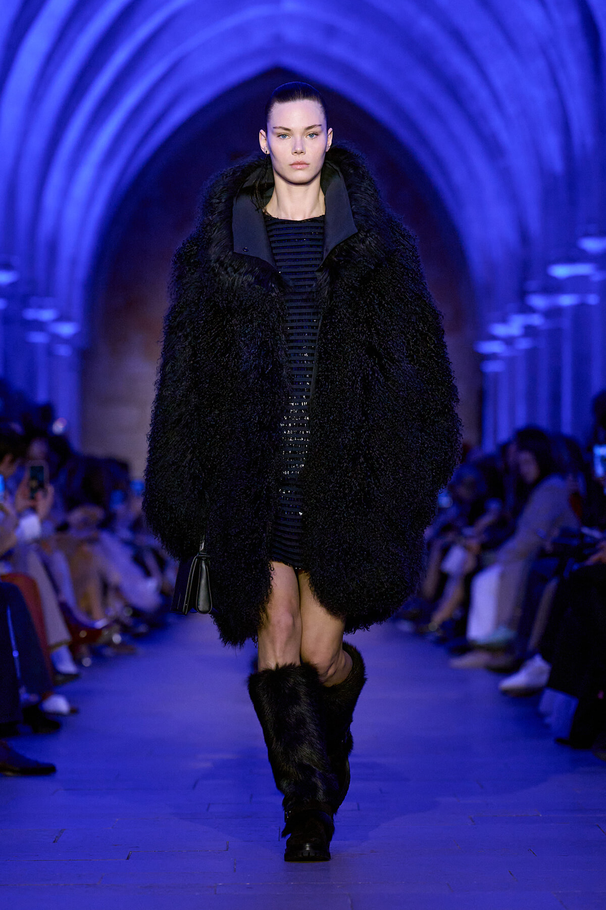 Model strides down a runway in a oversized black faux-fur coat, paired with fur boots, under blue-lit arches with an audience on sides