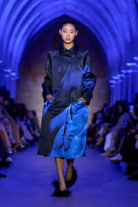 Model on a blue-lit runway wearing a glossy cobalt satin coat with hands in pockets.
