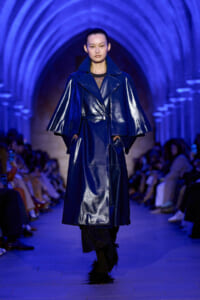 Model in a shiny cobalt trench coat with wide cape-like sleeves walks a blue-lit runway.