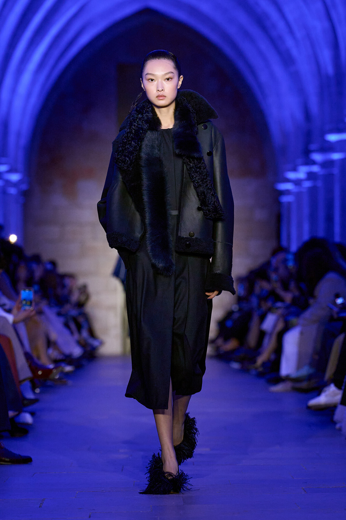 Model walking a fashion runway in a black fur-trimmed coat under blue lighting.
