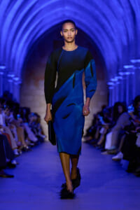 Model walking a blue-lit runway in a knee-length blue and black color-block dress, carrying a black clutch.