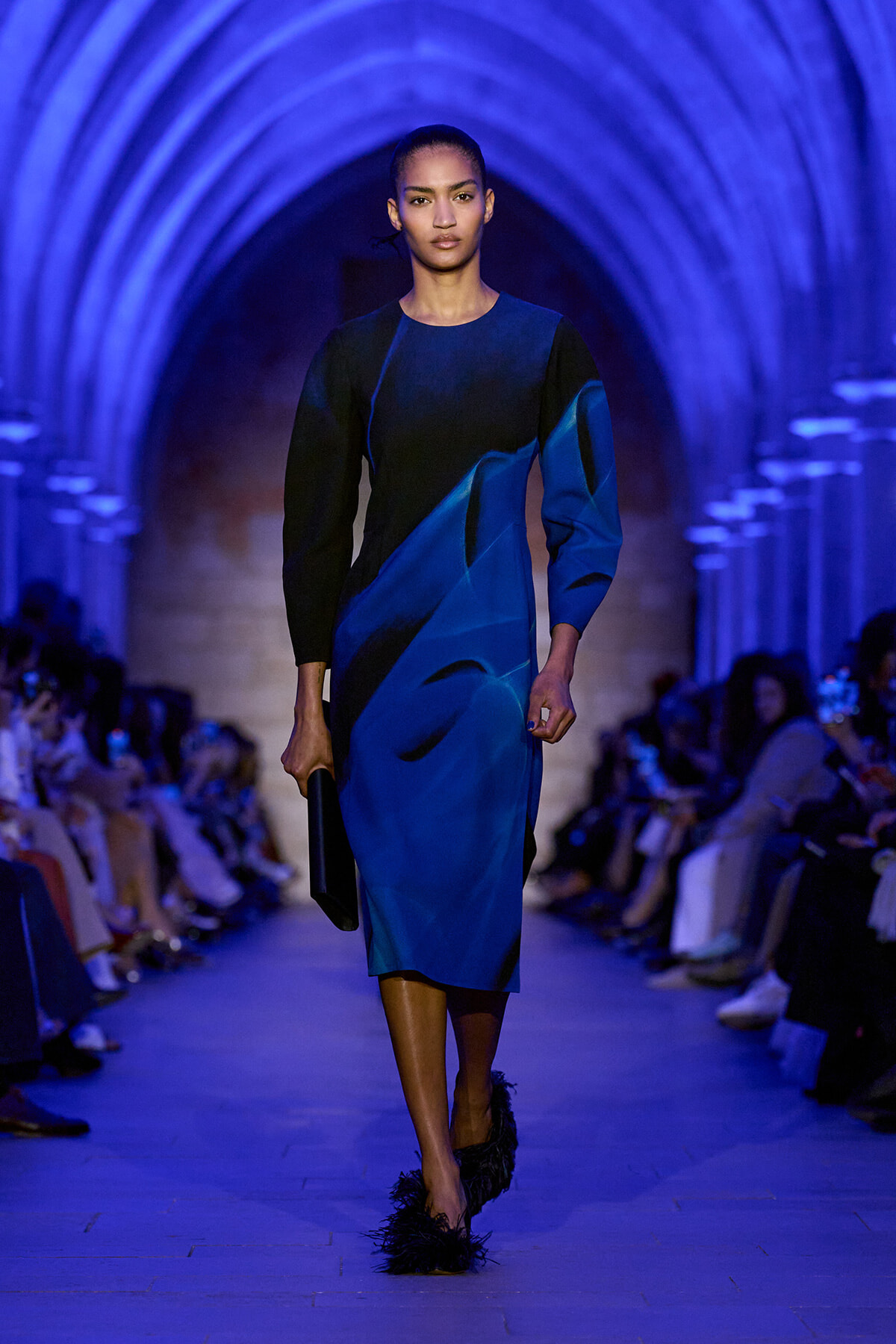 Model walking a blue-lit runway in a knee-length blue and black color-block dress, carrying a black clutch.