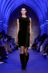 Model on a runway wearing a black asymmetrical dress with feather trim, blue arched backdrop and audience watching.