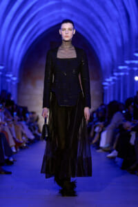 Female model walks a fashion runway in a black sequined top with a sheer neckline and a long, sheer black skirt under blue-lit arches.
