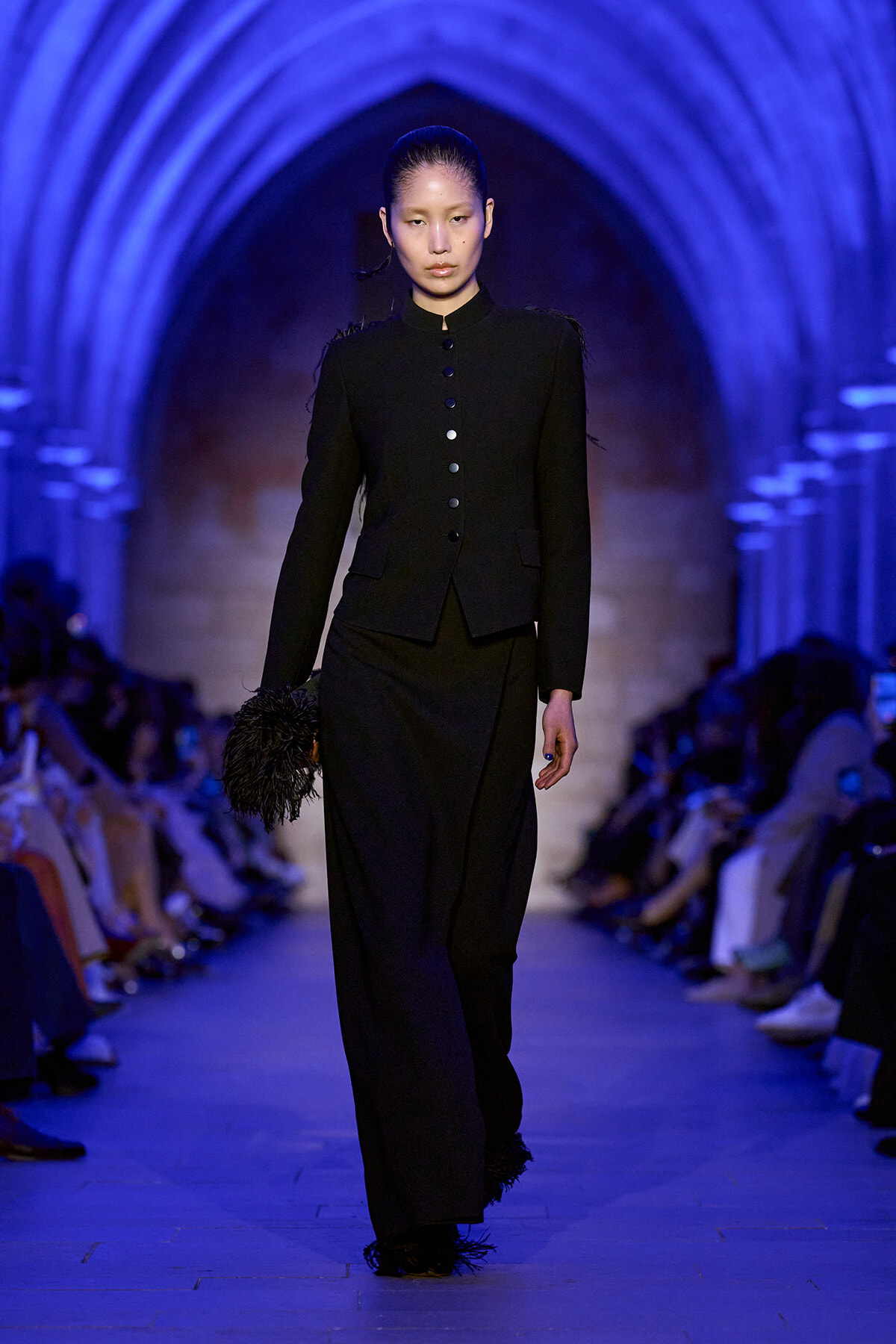Model walks a runway in a black tailored suit with a high collar and buttoned front, in a blue-lit archway backdrop.