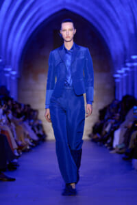 Model walking a runway in a cobalt blue suit with a matching shirt, under blue arched lighting at a fashion show.