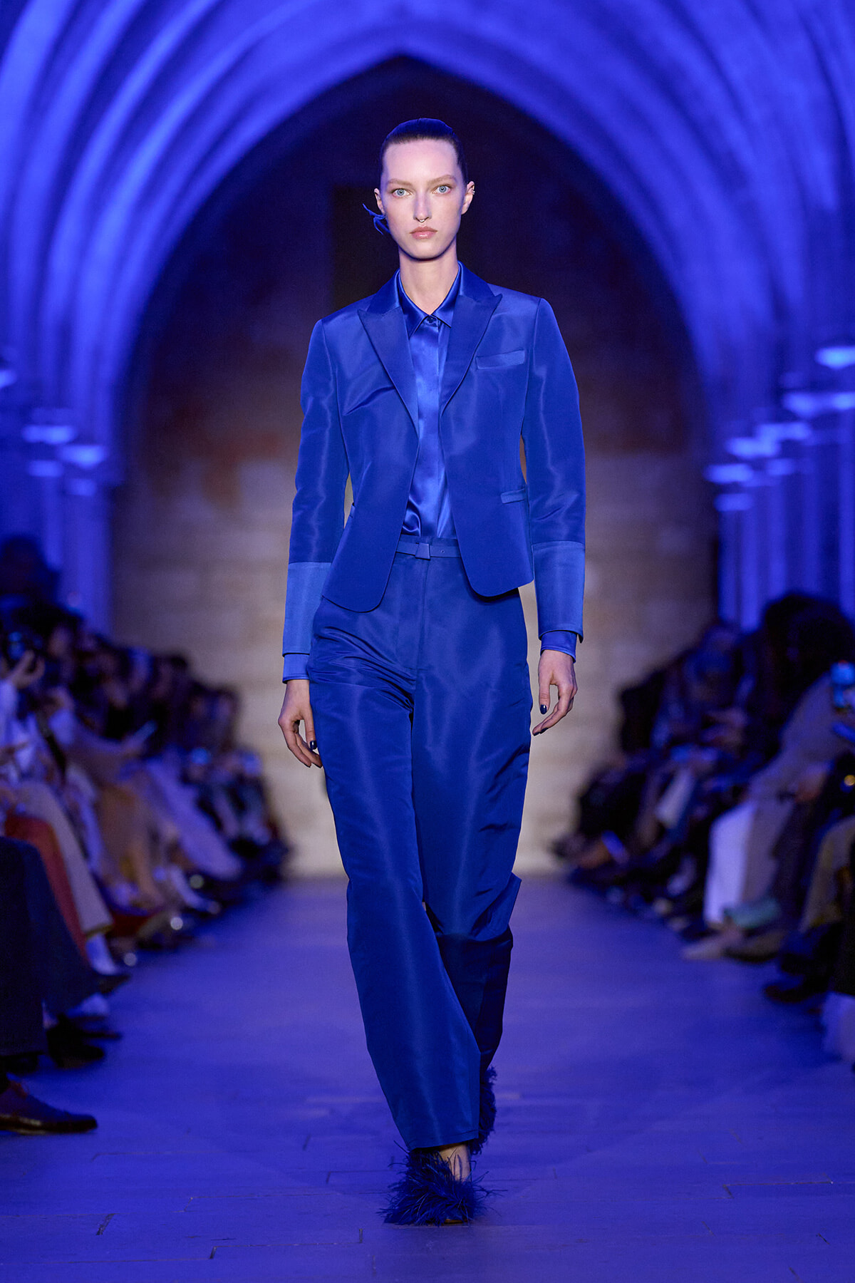 Model walking a runway in a cobalt blue suit with a matching shirt, under blue arched lighting at a fashion show.