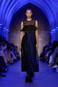 Model walks a runway in a sheer black top with feathered fringe and a navy-blue midi skirt under blue arches.