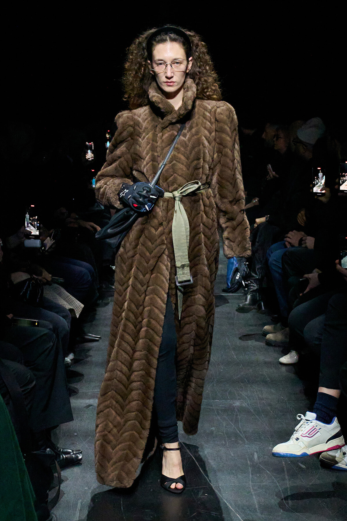 Model walks the runway in a long brown fur coat with a green belt, black gloves, and open-toed black heels.