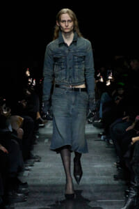 Model walks the runway in a blue denim jacket and matching denim skirt, with black tights and pointed heels, wearing glasses and gloves.