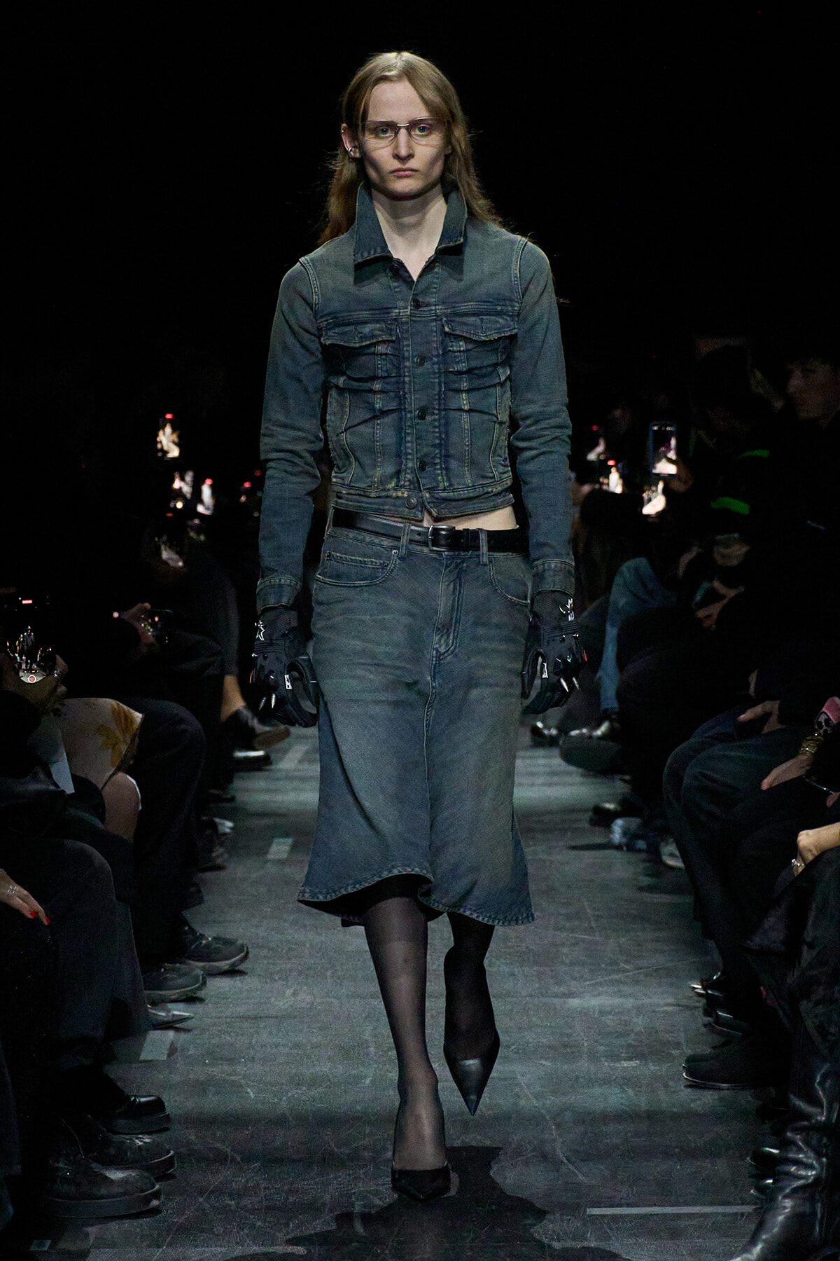 Model walks the runway in a blue denim jacket and matching denim skirt, with black tights and pointed heels, wearing glasses and gloves.