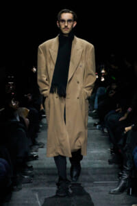 Male model walks the runway in a tan oversized double-breasted overcoat with a black scarf and sunglasses.