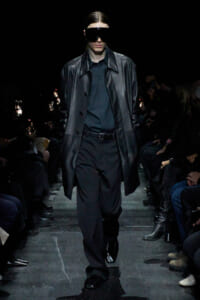 Model walking a runway in an all-black outfit: long leather-look coat over a black shirt and trousers, wearing oversized black sunglasses. Audience along the catwalk.