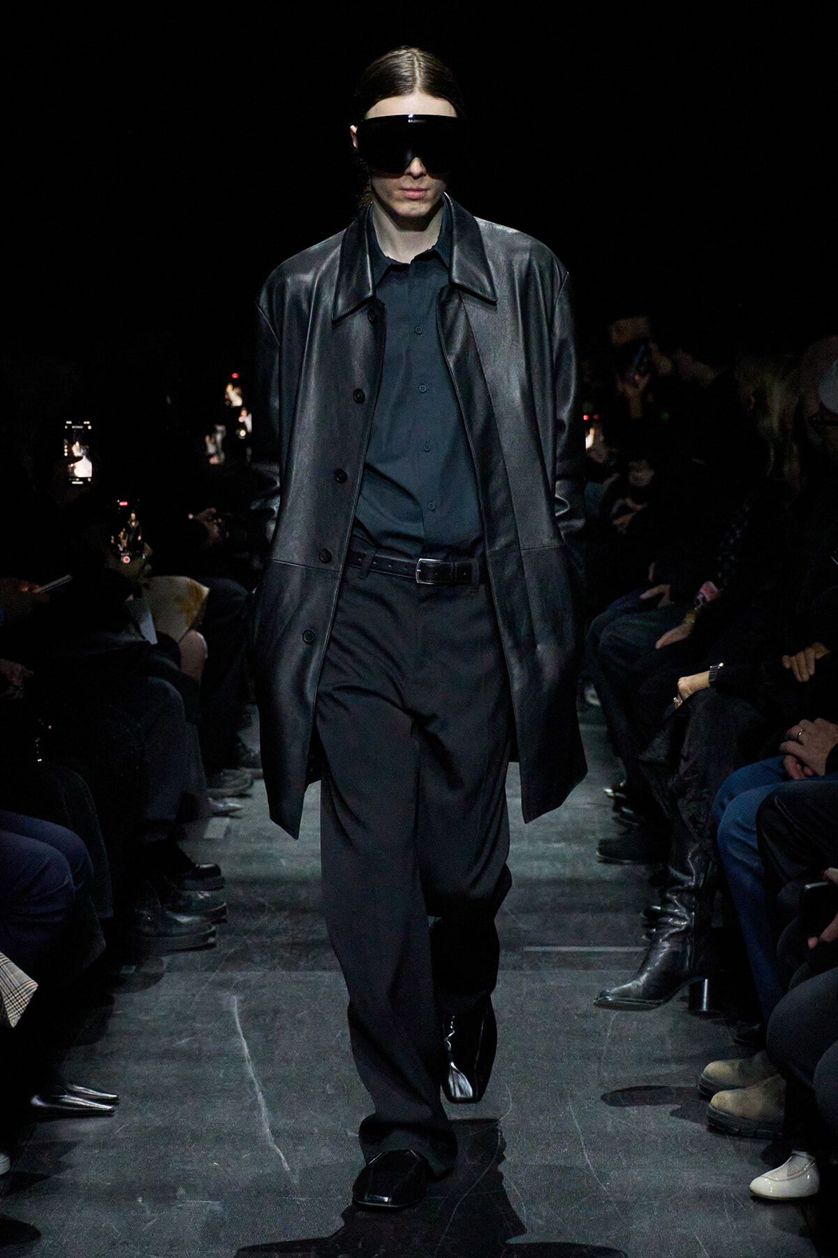 Model walking a runway in an all-black outfit: long leather-look coat over a black shirt and trousers, wearing oversized black sunglasses. Audience along the catwalk.