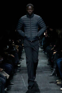 Male model walking a fashion runway in a dark quilted puffer jacket with hands in pockets, audience in shadows.
