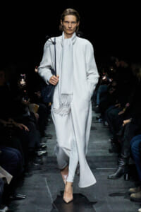 Model walks the runway in a long pale coat with a scarf, carrying a dark handbag, surrounded by seated audience in dim lighting.