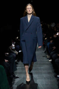Model walks the runway in a navy double-breasted coat with black heels, audience dimly lit on both sides of the catwalk.