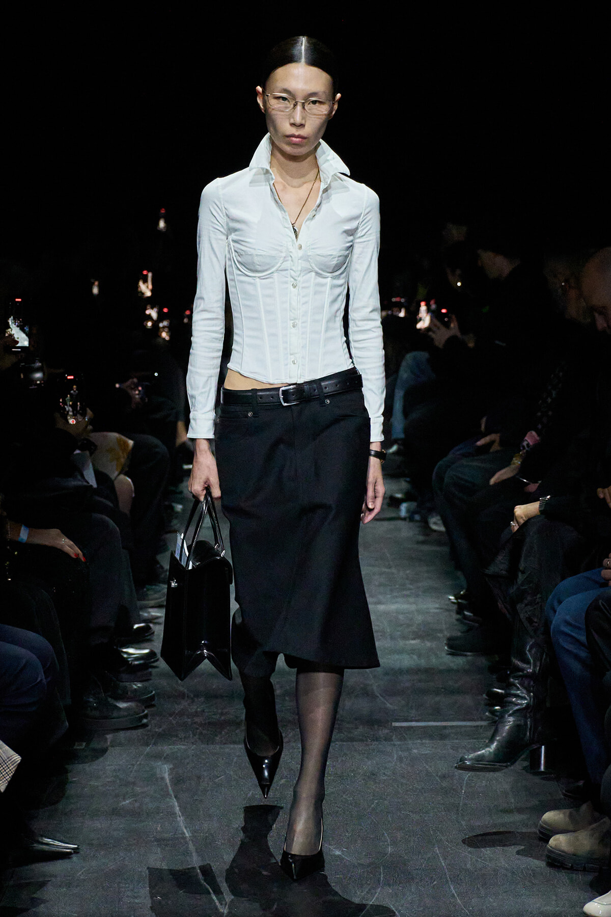 Model walking a fashion runway in a white fitted blouse with a corset waist and a black midi skirt, carrying a black handbag.