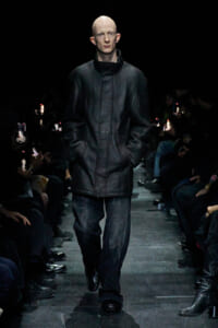 Male model walking a fashion runway in a black leather oversized jacket and dark denim pants, hands in pockets, audience filming with smartphones on either side.