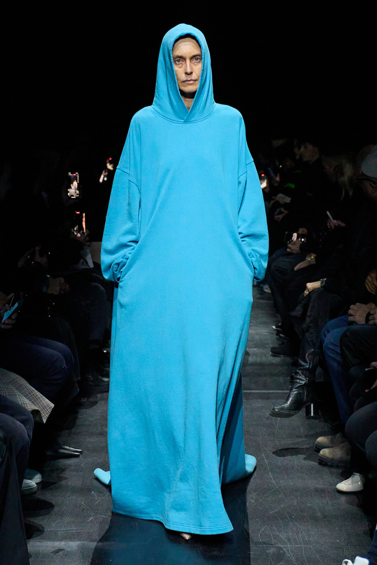 Model on a dim runway wearing a bright blue oversized hooded robe, hands in pockets, audience on both sides.