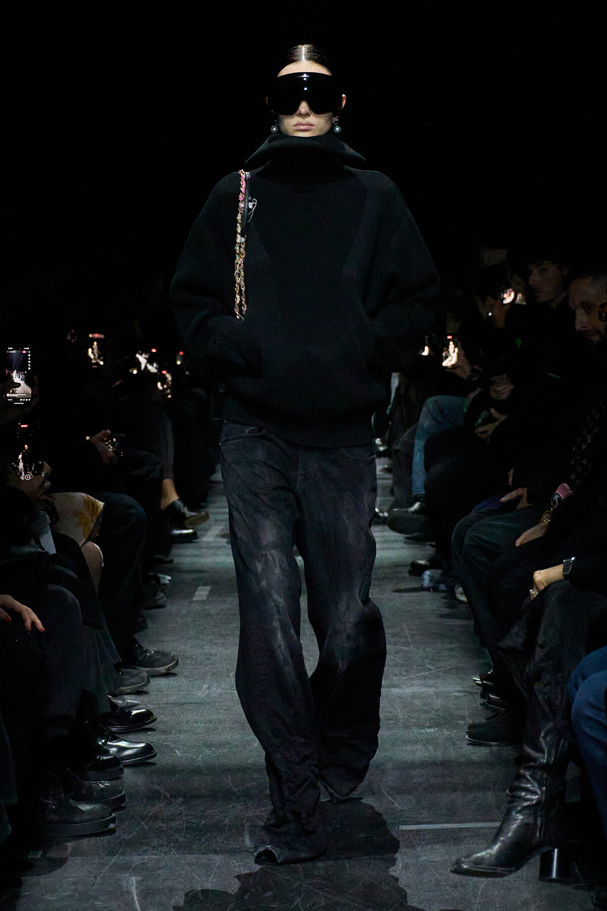 Model walks the runway in an oversized black sweater, wide dark pants, and large sunglasses; audience watches in the dark.