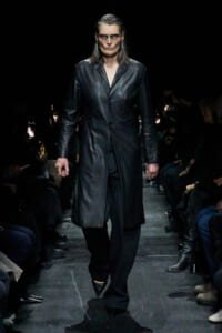 Model walks a long black leather trench coat down a dimly lit runway, audience on both sides.
