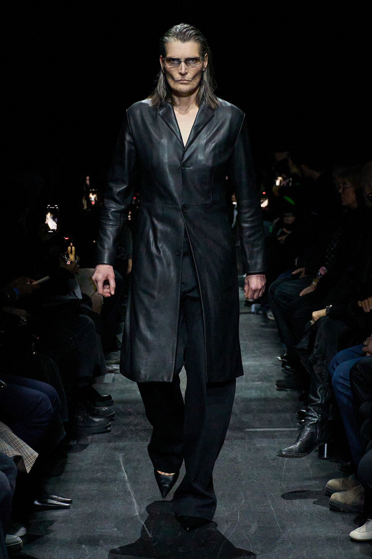 Model walks a long black leather trench coat down a dimly lit runway, audience on both sides.