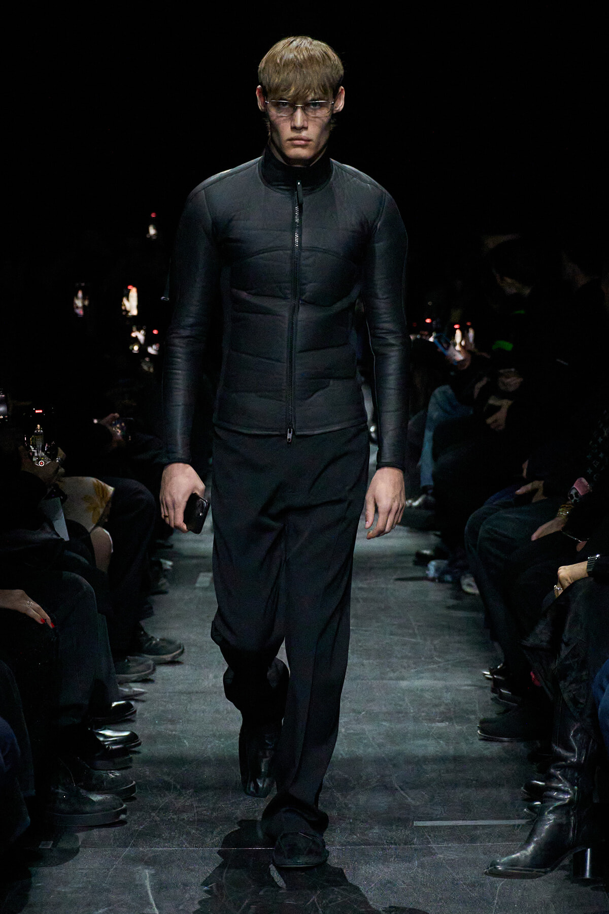 Male model walks a fashion runway in a black padded zip jacket and dark pants, audience in shadowed background.