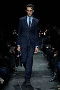 Male model in a navy suit and tie walks a runway, carrying a black briefcase, with spectators watching on both sides.