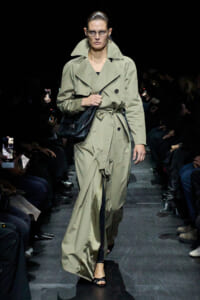 Model walking a fashion runway in a long beige trench coat with belt, carrying a black bag, wearing glasses and black sandals.