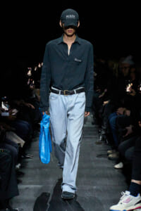 Male fashion model walking the runway in a dark button-down shirt, light blue jeans, a black belt, and a black cap reading 'MAIN'; he carries a bright blue bag in his left hand.