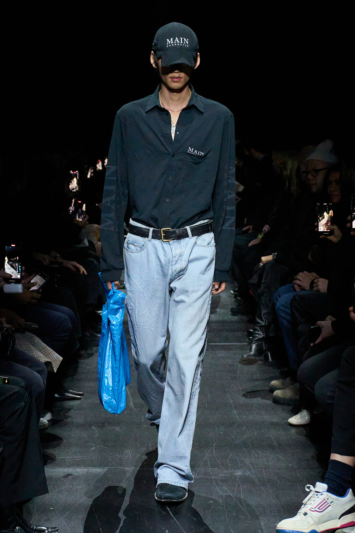 Male fashion model walking the runway in a dark button-down shirt, light blue jeans, a black belt, and a black cap reading 'MAIN'; he carries a bright blue bag in his left hand.