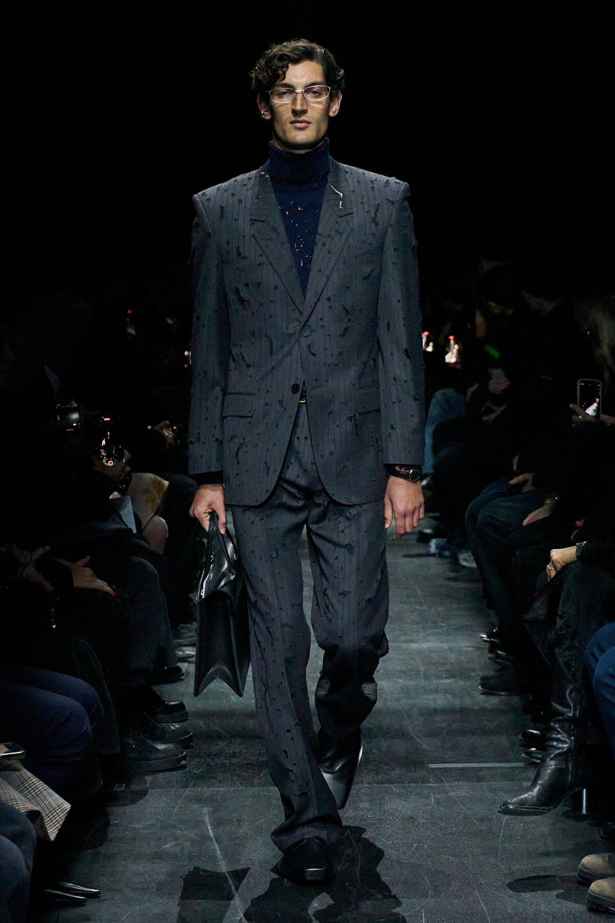 Male model walks the runway in a dark gray pinstripe suit with a navy turtleneck, carrying a black portfolio.