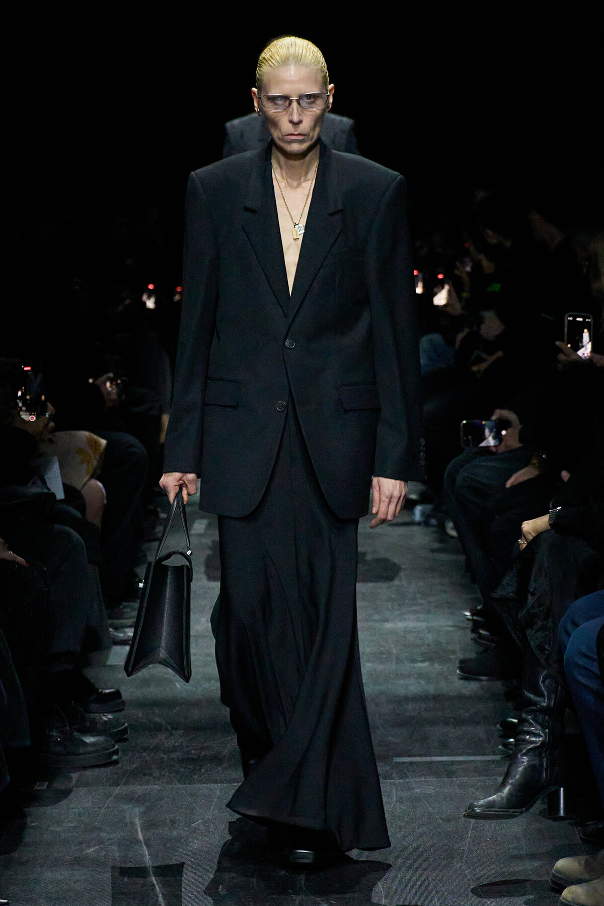 Model walking a runway in an oversized black suit, blonde slicked-back hair, sunglasses, gold pendant necklace, and carrying a black handbag.
