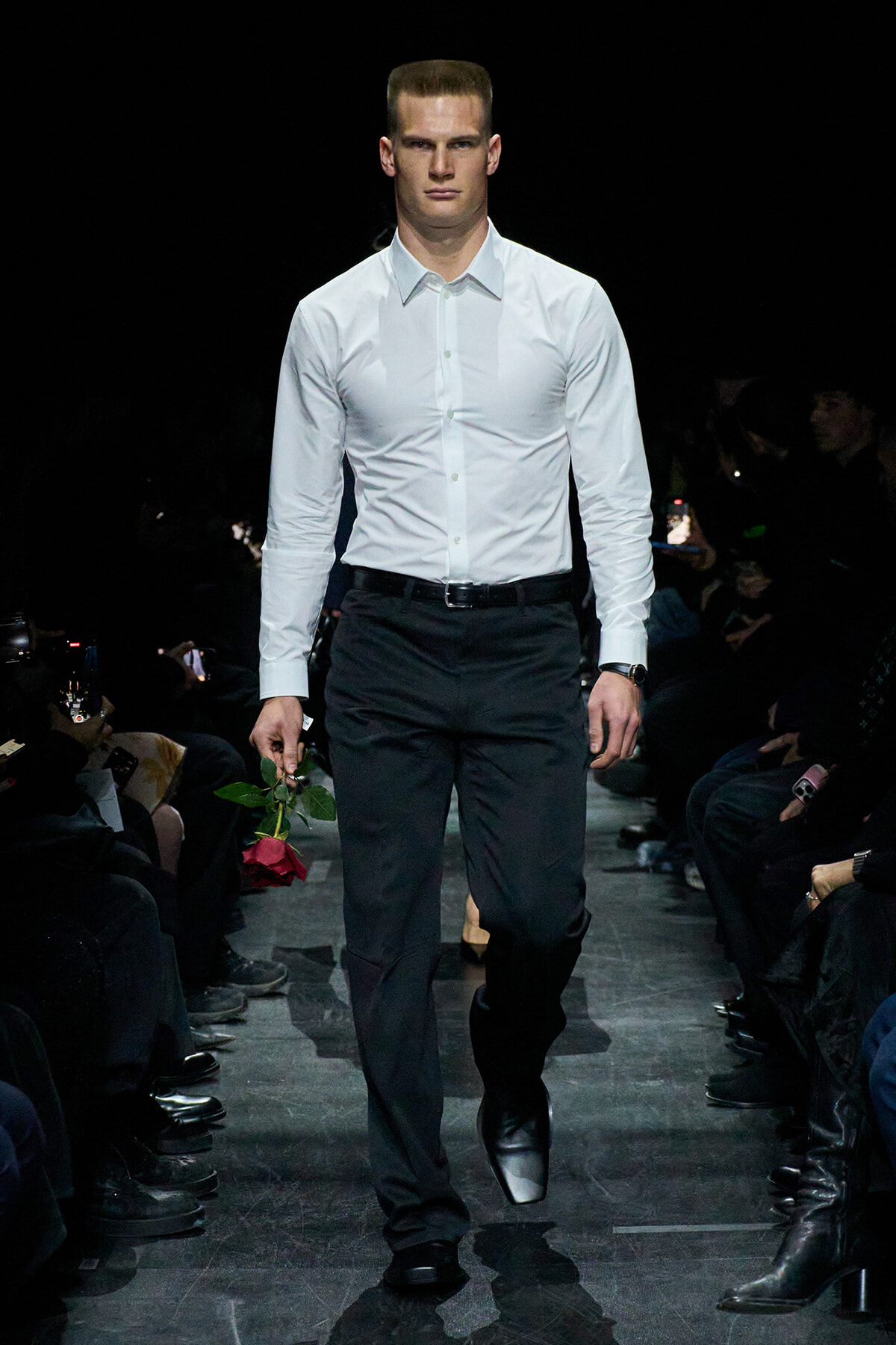 Male model walking a dim runway in a white dress shirt and black trousers, holding a red rose.