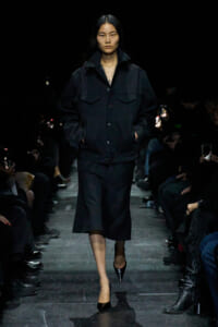 Model walks a dark runway in an all-black ensemble: oversized jacket over a knee-length skirt, hands in pockets, pointed heels.