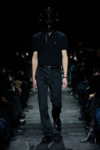Model walks a dark runway in an all-black outfit: polo shirt, tailored pants, and glossy full-face helmet obscuring the face.
