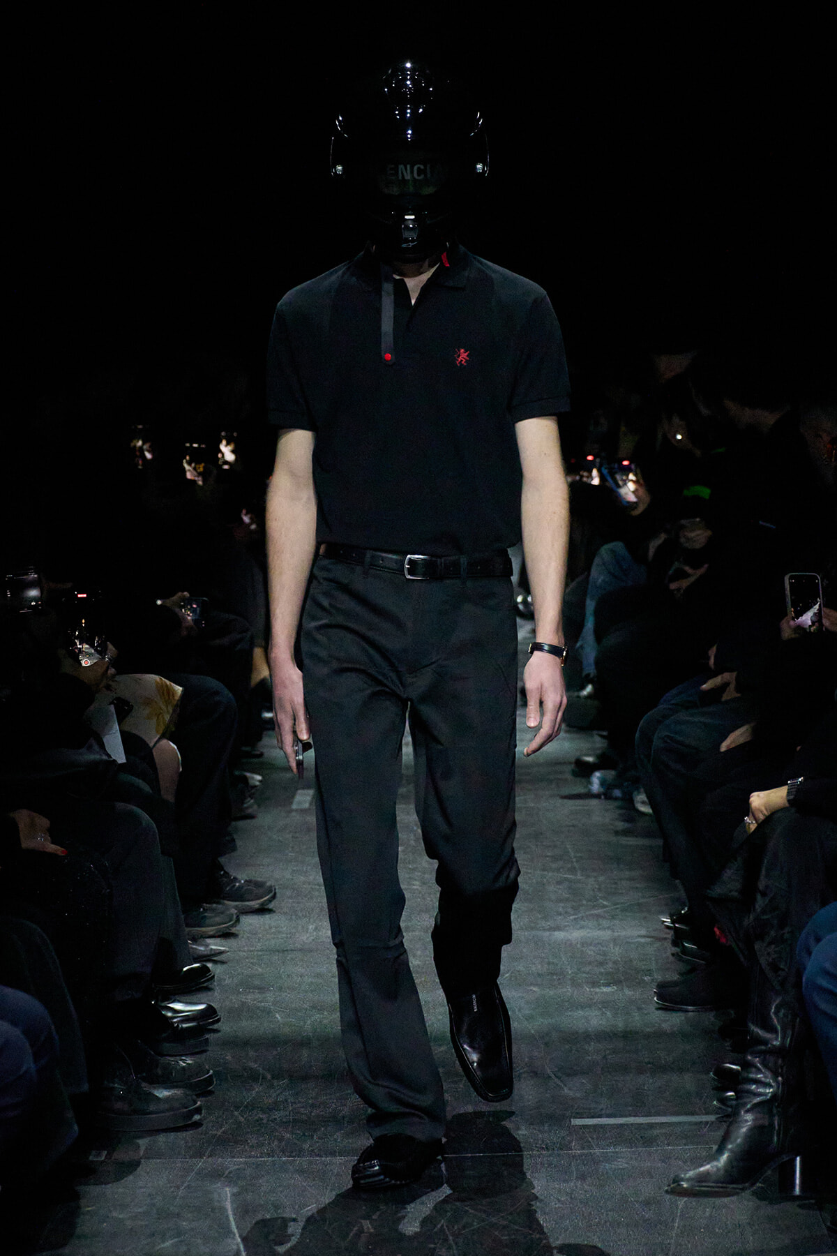 Model walks a dark runway in an all-black outfit: polo shirt, tailored pants, and glossy full-face helmet obscuring the face.