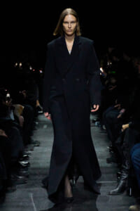 Model walks the runway in a long black double-breasted coat, minimal accessories, dark venue with seated audience on sides.
