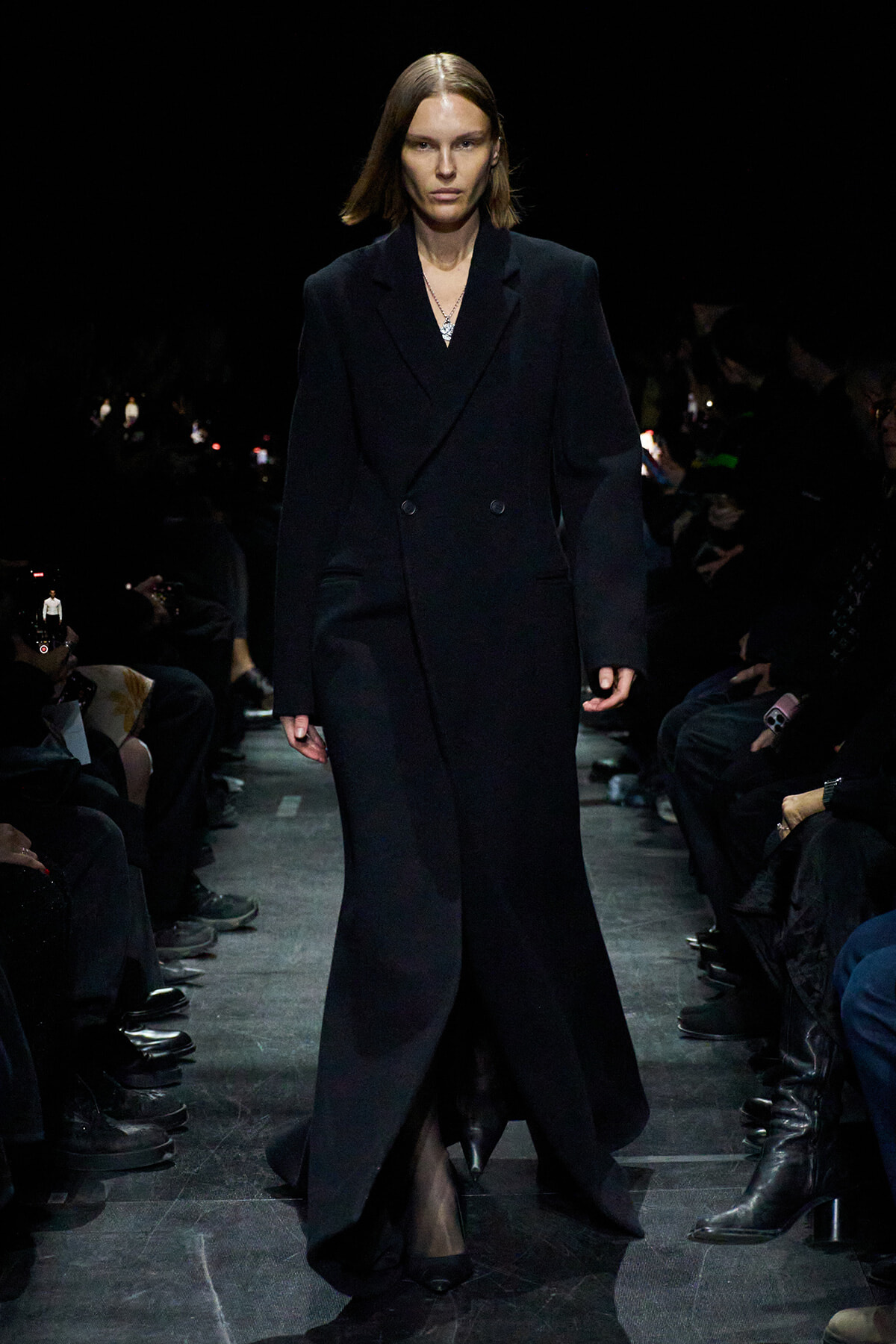 Model walks the runway in a long black double-breasted coat, minimal accessories, dark venue with seated audience on sides.