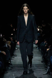 Model walks the runway in an oversized black suit with a deep V opening and black boots.