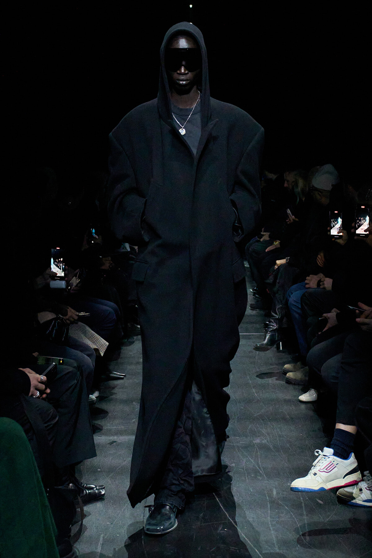 Model wearing an oversized black hooded coat and dark sunglasses walks a dim runway, audience on either side.