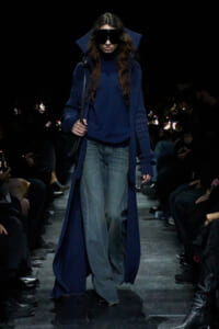 Model walking a dark runway in a long, navy blue coat over a matching sweater and wide-leg jeans, wearing oversized sunglasses.