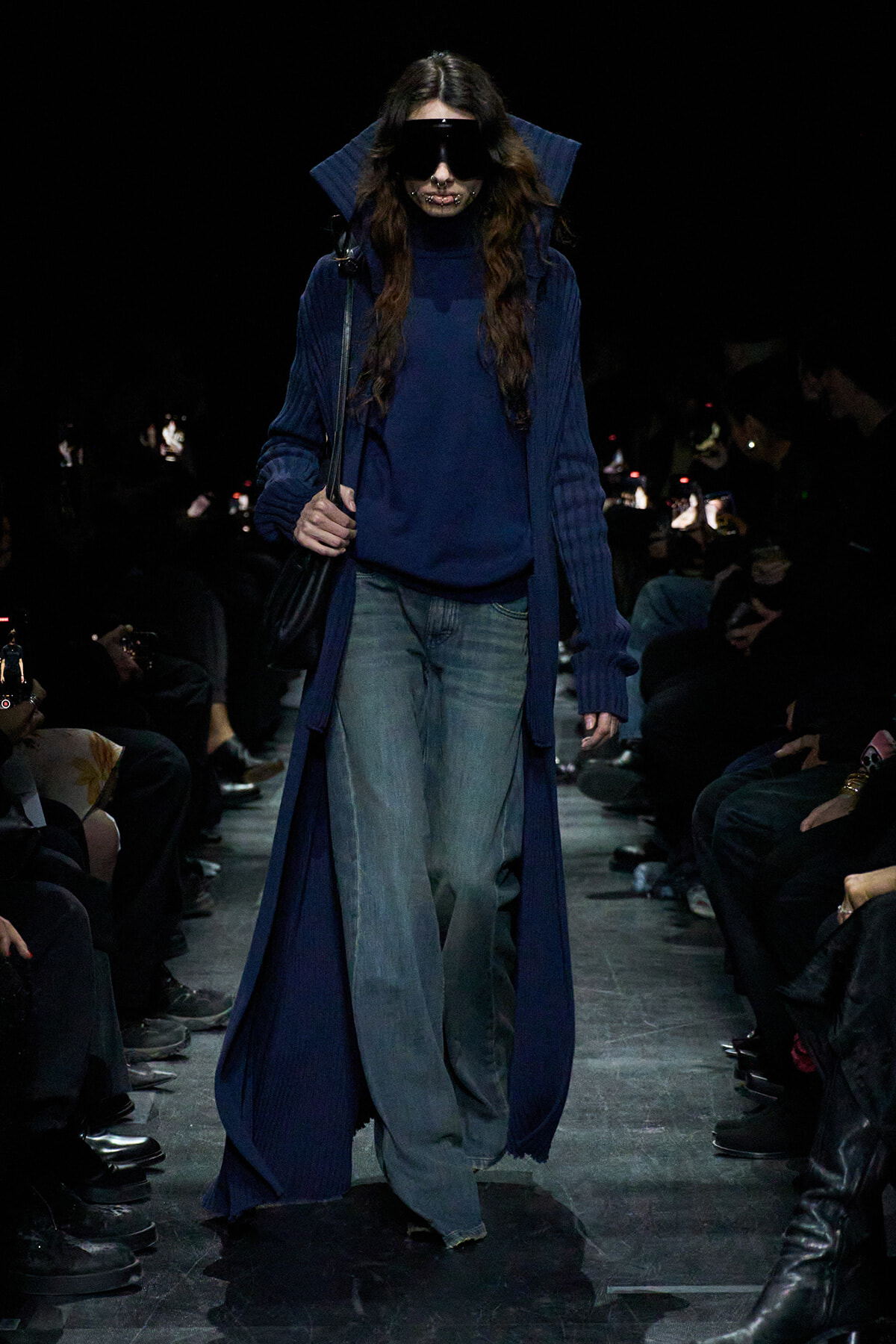 Model walking a dark runway in a long, navy blue coat over a matching sweater and wide-leg jeans, wearing oversized sunglasses.