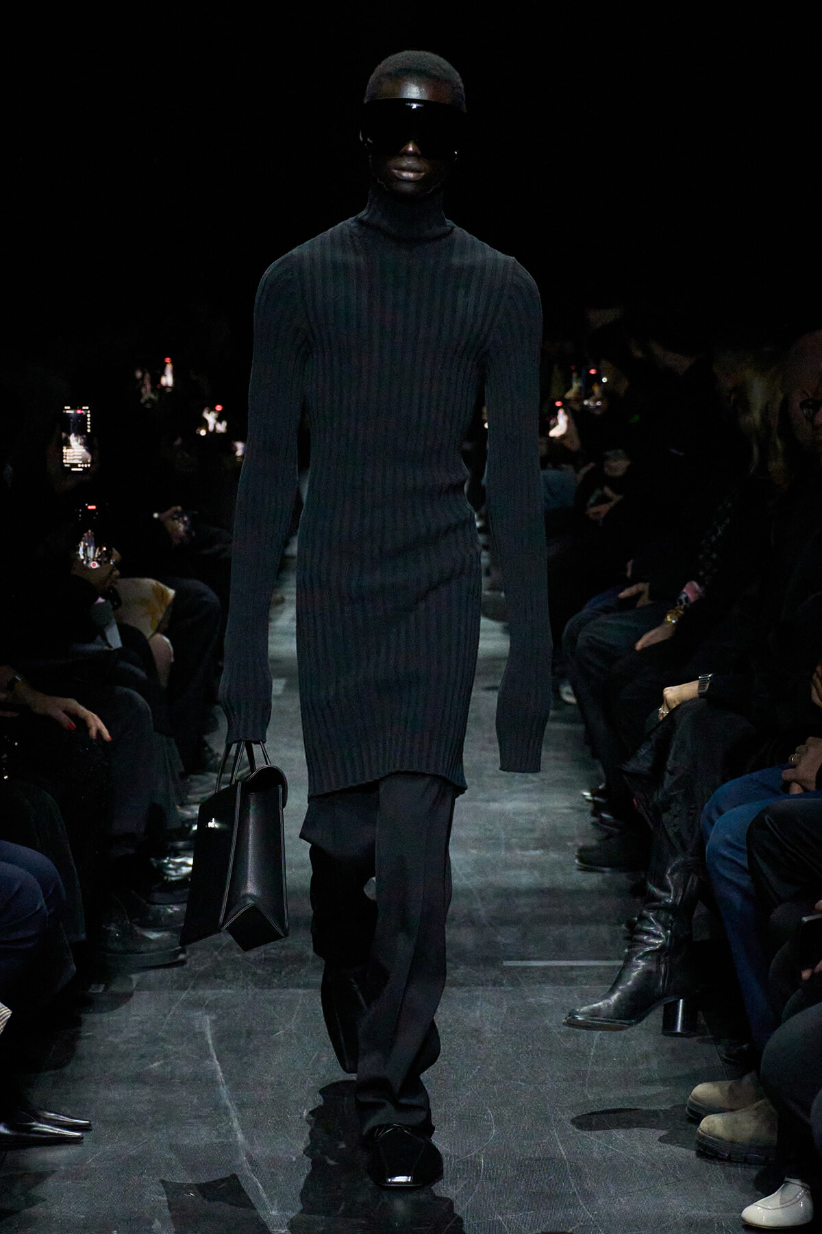 Model walking a fashion show in a black ribbed turtleneck dress with oversized sunglasses, carrying a black structured bag on a dark runway.