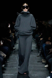 Model on a runway wearing a dark gray oversized knit top with a high collar and matching maxi skirt, plus large black visor sunglasses.
